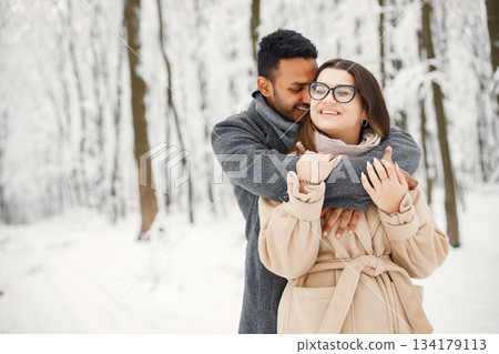 Indian man and caucasian woman walking in winter forest together. Lovely couple hugging. Man wearing grey coat and woman beige coat. Indian man and caucasian woman walking in winter forest together. Lovely couple hugging. Man wearing grey coat and woman beige coat. 134179113