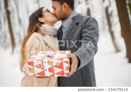 Indian man and caucasian woman walking in winter forest together. Man and woman holding a gift box. Man wearing grey coat and woman beige coat. Indian man and caucasian woman walking in winter forest together. Man and woman holding a gift box. Man wearing grey coat and woman beige coat. 134179114