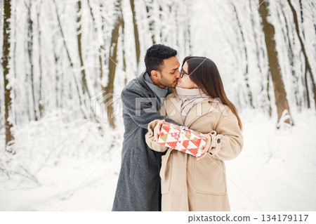 Indian man and caucasian woman walking in winter forest together. Man and woman holding a gift box. Man wearing grey coat and woman beige coat. 134179117