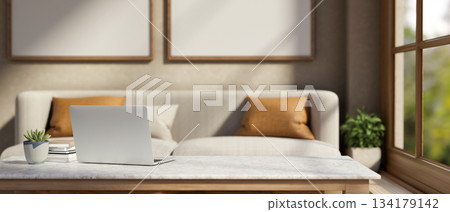 Laptop with book and pot plant on marble table across sofa with cushion aside window in living room. 134179142