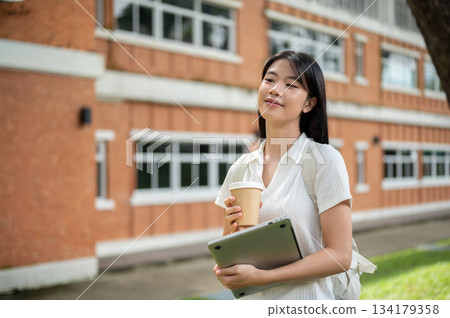 Young asian woman student holding laptop and coffee cup walking along brick wall building and garden Young asian woman student holding laptop and coffee cup walking along brick wall building and garden 134179358