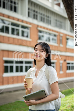 Young asian woman student holding laptop and coffee while walking by brick wall building and garden. Young asian woman student holding laptop and coffee while walking by brick wall building and garden. 134179359