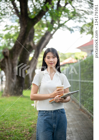 Young asian woman student holding coffee over book and laptop walking on garden sidewalk aside fence 134179361