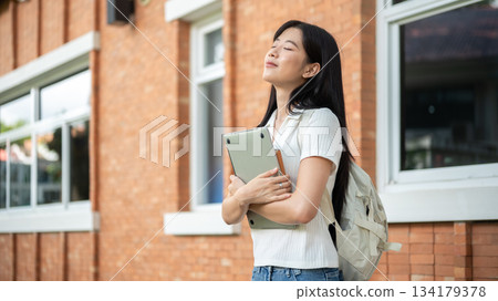 Asian woman student closing eyes holding hugging laptop and book standing aside brick wall building 134179378