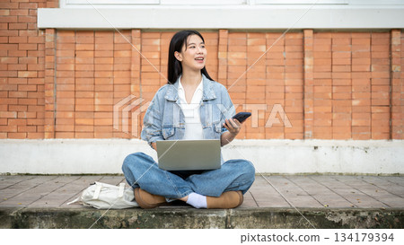 Young asian woman student holding phone over laptop looking away while sitting on street's sidewalk. Young asian woman student holding phone over laptop looking away while sitting on street's sidewalk. 134179394