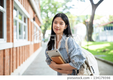 Young asian woman student with backpack holding hugging book and laptop on sidewalk around building. Young asian woman student with backpack holding hugging book and laptop on sidewalk around building. 134179398