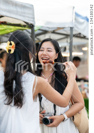 Asian woman feeding local food to her best friend who is standing holding a camera in local market. 134180142