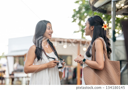 Pretty asian woman holding camera and talking with best friend while standing in the local market. Pretty asian woman holding camera and talking with best friend while standing in the local market. 134180145