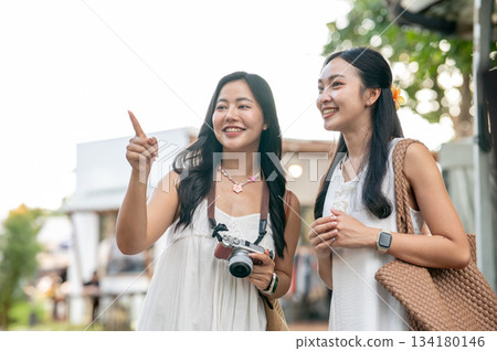 Pretty asian woman holding camera and pointing talking with a friend while standing in local market. Pretty asian woman holding camera and pointing talking with a friend while standing in local market. 134180146