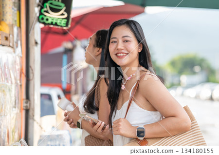 Pretty asian woman holding bag lookin away from phone while standing aside friend in a local market 134180155