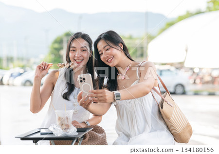 Pretty asian woman talking photo on phone with a friend holding eating food sitting in local market Pretty asian woman talking photo on phone with a friend holding eating food sitting in local market 134180156