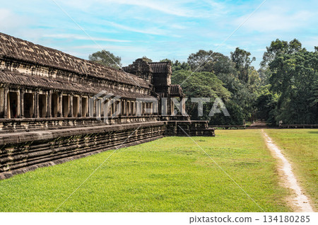 Wide angle view of massive stone wall and gallery of Angkor Wat temple with green grass field and footpath leading to forest 134180285