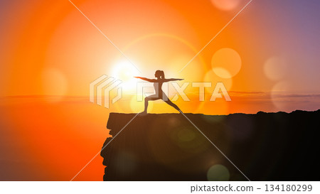 Silhouette of woman practicing yoga warrior pose on mountain cliff edge against orange sunset sky background illustrating freedom concept Silhouette of woman practicing yoga warrior pose on mountain cliff edge against orange sunset sky background illustrating freedom concept 134180299