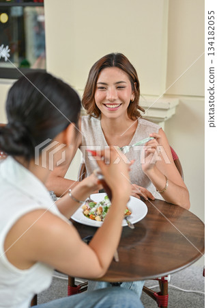 Asian woman holding fork talking with friend while sitting at wood table in front of restaurant cafe Asian woman holding fork talking with friend while sitting at wood table in front of restaurant cafe 134182055