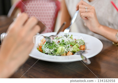 Close up woman's hands holding fork eating salad with friend sits at wooden table in restaurant cafe Close up woman's hands holding fork eating salad with friend sits at wooden table in restaurant cafe 134182058