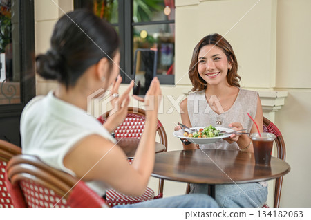 Asian woman holding phone taking picture of friend with salad plate at table in front of restaurant Asian woman holding phone taking picture of friend with salad plate at table in front of restaurant 134182063