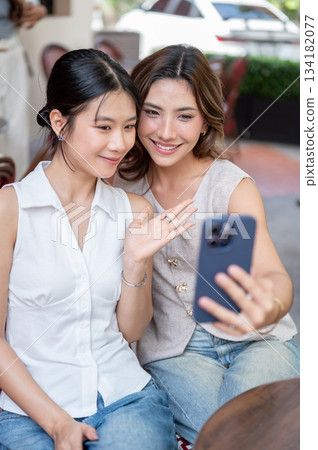 Pretty asian woman holding phone and taking picture with friend while sitting at table outside cafe Pretty asian woman holding phone and taking picture with friend while sitting at table outside cafe 134182077