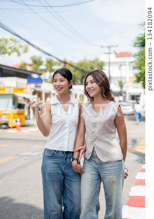 Pretty asian woman holding phone and pointing aside friend while standing or walking by street road. Pretty asian woman holding phone and pointing aside friend while standing or walking by street road. 134182094