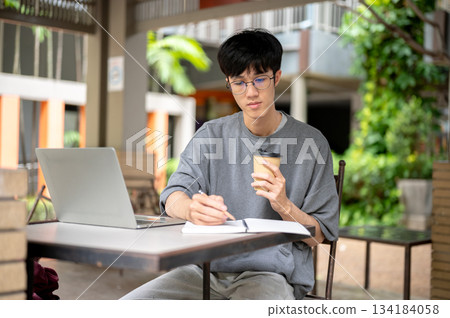 Glasses asian man holding coffee cup writing in notebook aside laptop while sitting at wooden table. Glasses asian man holding coffee cup writing in notebook aside laptop while sitting at wooden table. 134184058