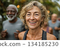 A joyful senior woman smiles brightly during an outdoor fitness activity, surrounded by friends jogging in the background, embodying health, energy, and vitality. 134185230