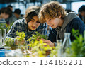 Two curious students closely examine plants in a bright science classroom, learning biology through hands-on experiments with greenery and lab glassware. 134185231
