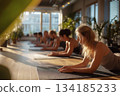 A serene yoga class practices on mats in warm sunrise light, with students aligned in a peaceful studio filled with plants and soft, calming atmosphere. 134185233
