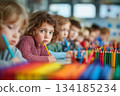 A young child with curly hair and bright blue eyes sits in a classroom, holding a colored pencil, surrounded by classmates and vibrant rows of crayons. 134185234