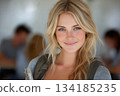 A smiling blonde student with bright blue eyes and a natural, fresh look stands confidently in a classroom setting, holding notebooks as soft light highlights her features. 134185235