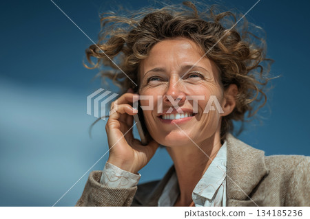 A cheerful woman smiles brightly while talking on her phone outdoors, her curly hair blowing in the wind under a clear blue sky, capturing a joyful, free moment. 134185236