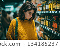 A young woman in a yellow sweater browses items in a grocery store aisle, appearing thoughtful and focused while shopping under soft, moody lighting. 134185237