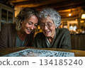A joyful moment between a senior woman and her adult daughter as they lean together over a puzzle, sharing smiles and warmth in a cozy, softly lit home setting. 134185240