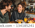 A young couple enjoys a cozy breakfast together, smiling warmly at each other in a relaxed kitchen setting filled with pastries, fruit, and fresh orange juice. 134185241