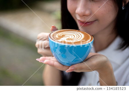 Close up of pretty asian woman holding latte art coffee in blue ceramic cup and smelling the aroma. 134185523