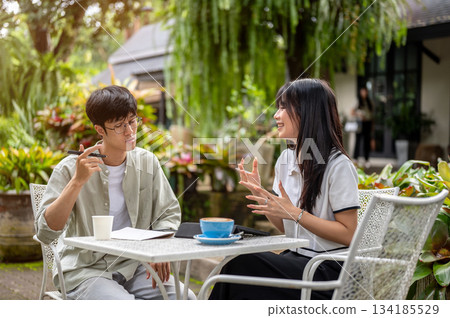 Glasses asian man and woman student friend talking while doing assignment at table in cafe's garden 134185529