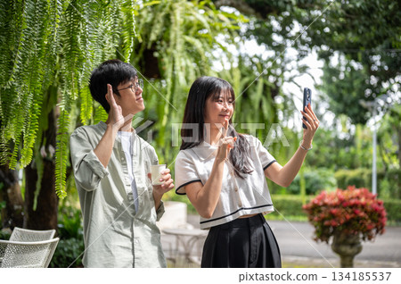 Pretty asian woman holding phone taking picture with glasses man standing under tree in cafe garden 134185537