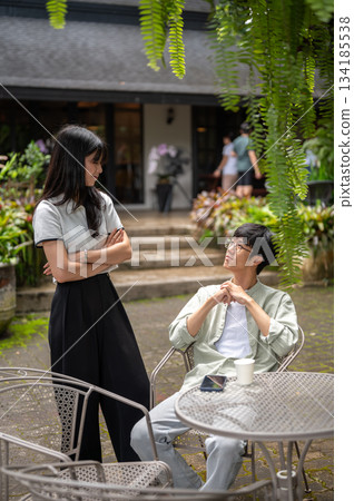Pretty asian woman standing with crossing arms looking at a glasses man sits at table in cafe garden 134185538