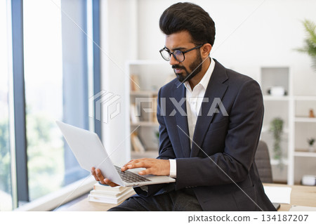 An Indian businessman wearing glasses works on his laptop in a modern office setting 134187207