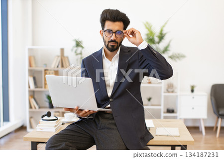 A stylish Arabian businessman is holding a laptop while wearing glasses and a suit 134187231