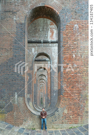 View of a young man wearing a black T-shirt and jeans standing on the structure under the bridge of Ouse Valley Viaduct or Balcombe Viaduct (Railway viaduct). View of a young man wearing a black T-shirt and jeans standing on the structure under the bridge of Ouse Valley Viaduct or Balcombe Viaduct (Railway viaduct). 134187405