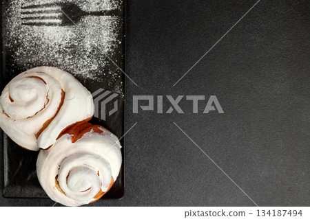 Fragrant cinnabons with cream on a black rectangular plate on a gray background Fragrant cinnabons with cream on a black rectangular plate on a gray background 134187494