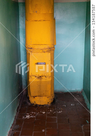 Inside an old building, a yellow garbage chute stands against a wall. The surrounding walls are in a faded blue, and the floor shows signs of wear and dirt 134187587