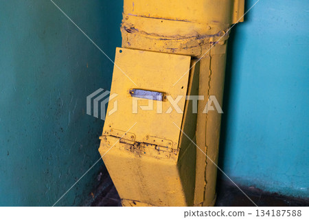 The yellow garbage chute in the corner of an old building has visible wear and rust. It stands against a faded blue wall, showing the building's age and previous usage The yellow garbage chute in the corner of an old building has visible wear and rust. It stands against a faded blue wall, showing the building's age and previous usage 134187588