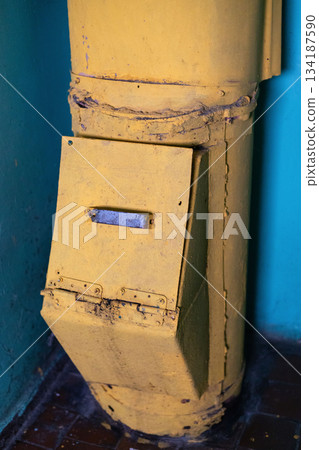 A weathered garbage chute stands in the corner of an old, neglected building. The bright yellow surface is scratched and rusty, revealing the passage of time and use 134187590