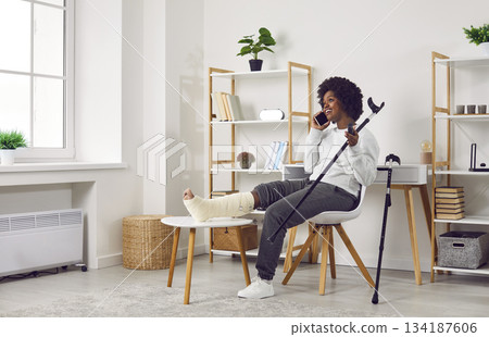Young woman with injured leg sitting at home on chair with crutches and talking on the phone. Young woman with injured leg sitting at home on chair with crutches and talking on the phone. 134187606