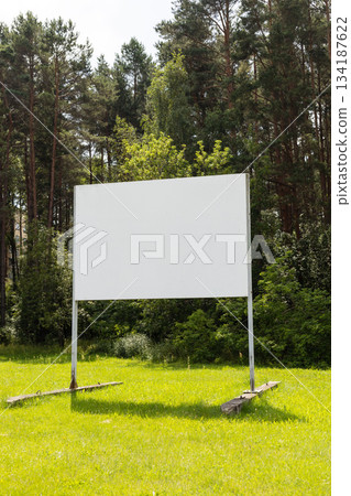 A large blank billboard is placed in the middle of a vibrant green field, bordered by tall trees. The scene is serene and peaceful, suggesting potential for advertisement or information A large blank billboard is placed in the middle of a vibrant green field, bordered by tall trees. The scene is serene and peaceful, suggesting potential for advertisement or information 134187622