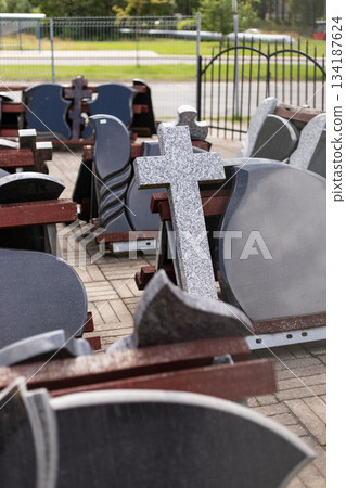 A variety of tombstones are set up for sale outdoors, showcasing different designs, materials, and styles. Customers browse the selection in a spacious area surrounded by greenery 134187624