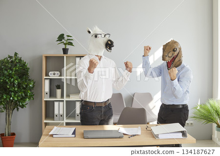 Two businessmen in dinosaur and horse animal masks raising their hands up in office Two businessmen in dinosaur and horse animal masks raising their hands up in office 134187627
