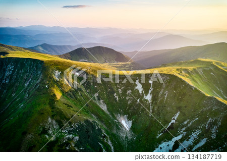 Aerial view of rugged mountain range with lush green slopes at sunset. Landscape showcases beauty of untouched nature with distant peaks and valleys bathed in soft sunlight. Carpathians, Chornogora. Aerial view of rugged mountain range with lush green slopes at sunset. Landscape showcases beauty of untouched nature with distant peaks and valleys bathed in soft sunlight. Carpathians, Chornogora. 134187719