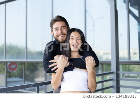 Happy young couple embracing in a loving pose near a glass window Happy young couple embracing in a loving pose near a glass window 134188315