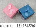 Two small boxes with ribbons are placed on a shiny surface. One box is blue and the other is pink. These boxes are ready for gifting during a party or event. 134188596
